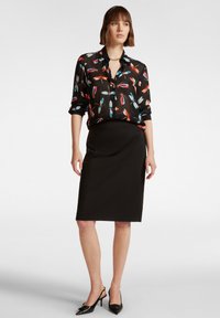 Camicia nera con motivo di piume colorate, maniche lunghe arrotolate, abbinata a una gonna a tubino nera aderente e décolleté nere a punta.