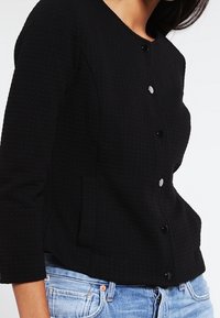 Veste noire texturée avec un col rond, une fermeture boutonnée visible et des poches latérales, associée à un denim bleu clair.