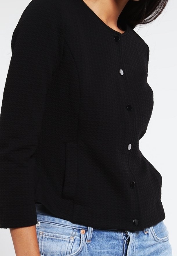 Veste noire texturée avec un col rond, une fermeture boutonnée visible et des poches latérales, associée à un denim bleu clair.