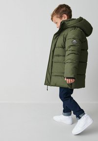 Giacca a vento verde con cappuccio, dotata di un patch a forma di diamante sulla manica sinistra, indossata sopra jeans scuri e sneaker bianche.