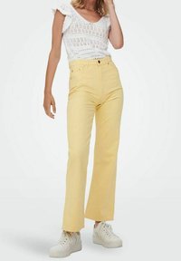 Żółte dżinsy z wysokim stanem i prostym krojem, o gładkiej fakturze i przednich kieszeniach, w zestawie z białymi trampkami i koronkową bluzką.