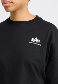 Juodas medvilninis sportinis megztinis su baltu "Alpha Industries" logotipu ir stilizuota emblema kairėje krūtinės pusėje. Apvalus iškirpimas ir laisvas kirpimas.