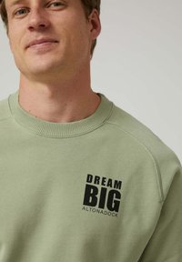 Hombre vestido con un sudadera verde claro con el texto negro "DREAM BIG ALTONADOCK" en la parte superior del pecho, fondo neutro.