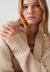 Beige gestrickter Cardi mit geripptem Kragen und Ärmeln. Weiche Textur, über einem braunen Oberteil getragen, verfügt über subtile Details und eine lockere Passform.