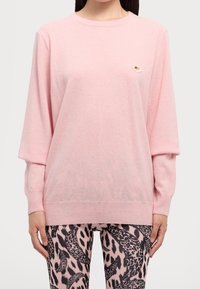 Pull rose clair avec un col rond et des manches longues. Présente un petit élément décoratif sur la poitrine. Porté avec des leggings à motifs.