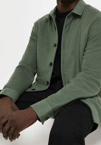 Zielona polarowa koszula-kurtka z guzikiem z przodu, kołnierzem typu spread oraz długimi rękawami. Gładka tekstura i luźny krój. Nosi się ją na czarnym topie.