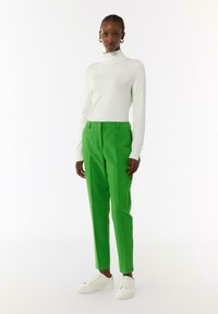 Pantaloni su misura verdi a vita alta, gamba dritta e passanti per cintura, abbinati a un dolcevita bianco attillato. Le sneakers bianche completano il look.
