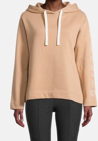 Beige hoodie med vita dragkedjor och text på ärmen. Har en avslappnad passform och mjukt tyg, lämplig för avslappnad klädsel.