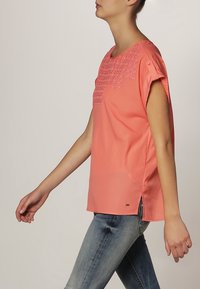 Koralfarvet bluse med korte ærmer, et tekstureret blomstermønster ved halsen og sideslids. Båret med slim-fit jeans.