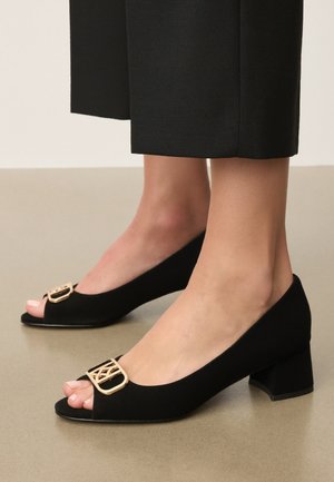 Black low-heeled peep-toe shoes with gold decorative buckle worn with black cropped pants on bare feet.