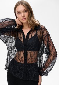 Czarna koronkowa bluzka z długimi, przezroczystymi rękawami, z zapięciem na guziki z przodu oraz jednolitą czarną kołnierzykiem. Kwieciste wzory na całej powierzchni.