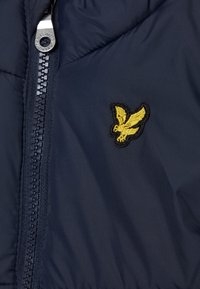 Granatowa kurtka puchowa z zapięciem na suwak ma haftowane żółte logo ptaka po lewej stronie klatki piersiowej oraz gładką, teksturowaną tkaninę.