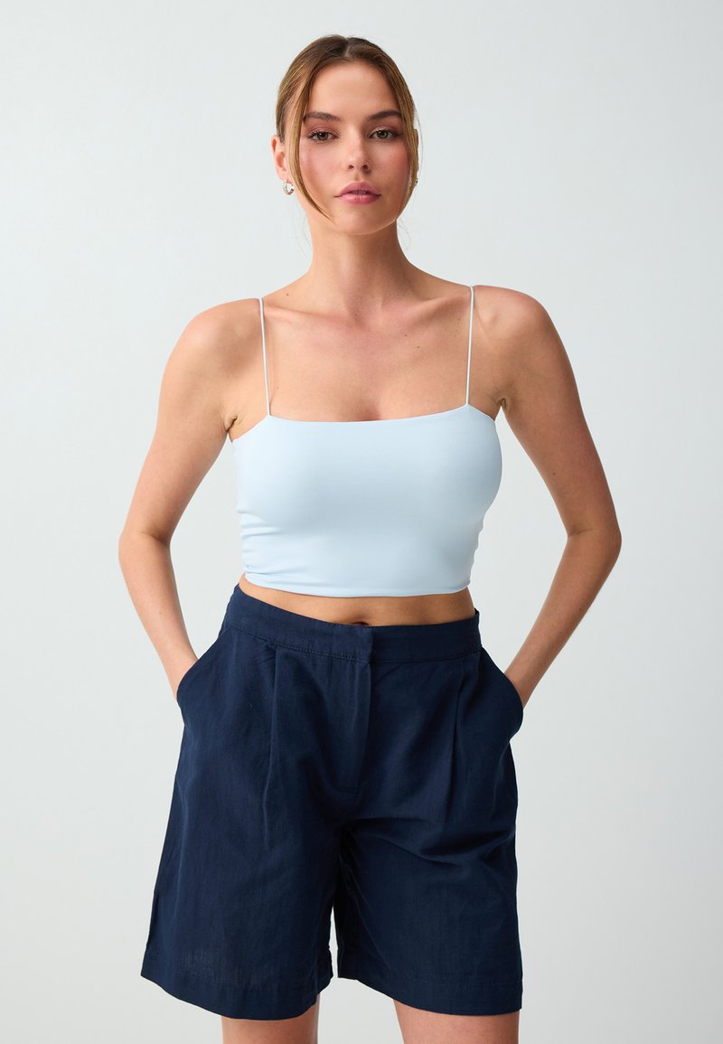 Femme portant un crop top bleu clair à fines bretelles et un short bleu marine taille haute, debout les mains dans les poches.