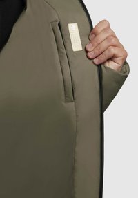 Oliivinvihreä takki, jossa on sileä pinta; siinä on vetoketjukiinnitys ja pieni beige etiketti sisätaskun yläpuolella.