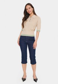 Beige ribbed polo top with three buttons, short sleeves, and green striped accent at the hem, paired with dark blue cropped jeans and black heels.
