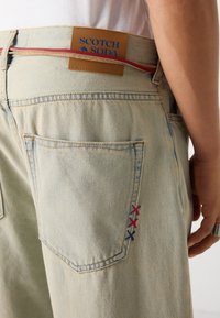 Light blue denim jeans featuring a leather label with blue text. Notable red and blue stitching on the back pocket adds detail.