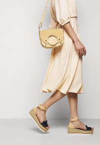 Gul mockaskinnväska med cirkulär detalj och guldhardware, bärs med en ljusbeige klänning och tan espadriller med marinblå accenter.