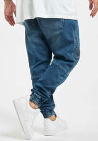 Niebieskie dżinsowe joggery z elastycznymi ściągaczami, wyposażone w teksturowany boczny panel i standardowe tylne kieszenie, zestawione z białymi trampkami.