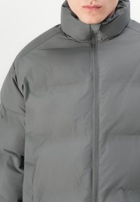 Hallhalline puffer jope kõrge kraega, täispikk lukk, tikitud tekstuur ja sujuvad õlavarred. Ei ole nähtavat riistvara ega logosid.