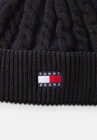 Gorro de malha preto com um design de malha em cabos e uma bainha canelada. Inclui um patch retangular com o texto "TOMMY JEANS" e as cores da bandeira.