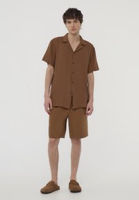 Camicia marrone a maniche corte con bottoni e colletto, abbinata a pantaloni corti coordinati. Il tessuto sembra leggero e leggermente strutturato.