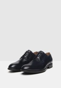 Scarpe da uomo in pelle blu navy con texture liscia, punta arrotondata e chiusura con lacci. Presentano un tacco basso e dettagli di cucitura minimi.