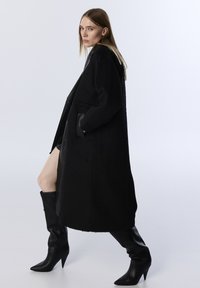 Schwarzer Wollmischungs-Mantel mit einem schlanken Design, ausgestattet mit großen Taschen und Lederelementen, kombiniert mit hohen, spitzen schwarzen Lederstiefeln.