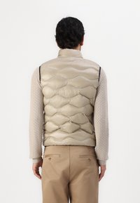 Gilet puffa trapuntato beige chiaro con colletto alto, caratterizzato da una texture lucida e da cuciture curve. Indossato sopra un maglione testurizzato e pantaloni.