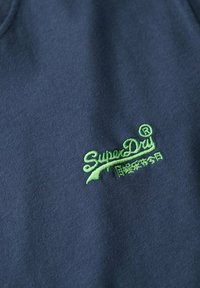 Un tessuto blu navy presenta un logo "SuperDry" ricamato in verde con un font stilizzato, caratterizzato da una superficie liscia e texture.