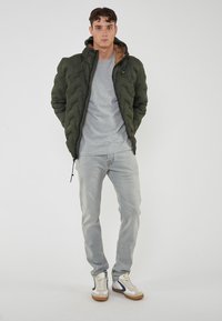 Giubbotto verde imbottito con zip frontale e fodera marrone, indossato sopra una maglietta grigia, abbinato a jeans grigi chiari e sneakers multicolori.