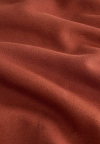 Tessuto lavorato a maglia color ruggine morbido, con pieghe e ombre sottili che evidenziano la sua trama e la superficie liscia.