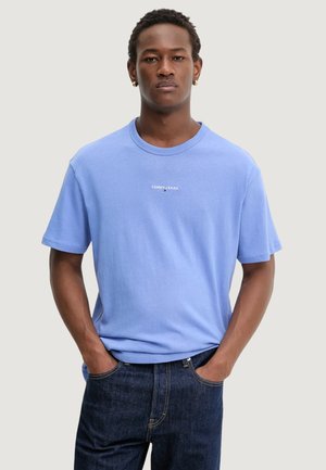 Giovane uomo con capelli corti e ricci che indossa una T-shirt Tommy Jeans azzurra e jeans blu scuro, mani in tasca, sfondo neutro.