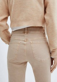 Un jean taille haute beige présente une coupe ajustée, cinq poches et des détails de couture subtils à l'arrière. Associé à un pull beige clair.