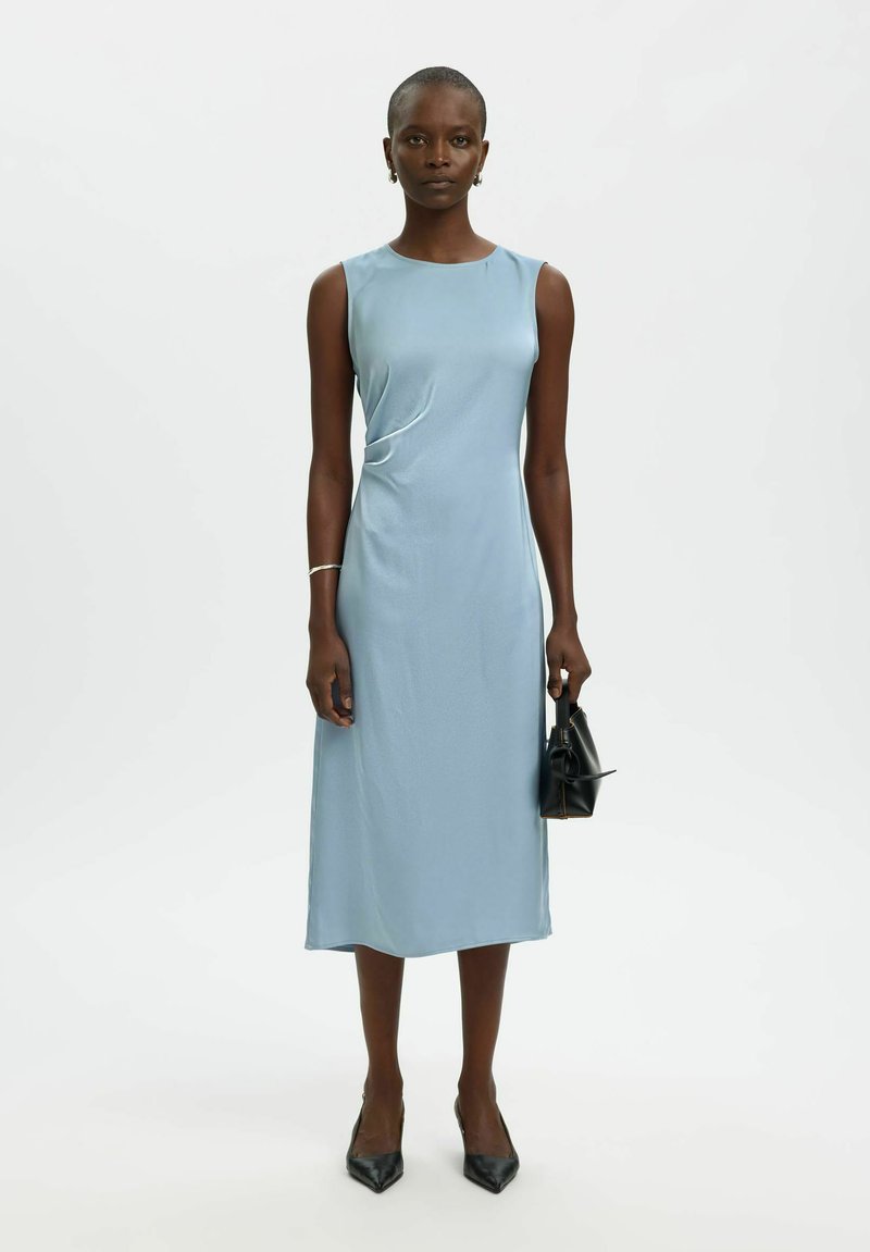 Femme portant une robe midi bleu clair sans manches, tenant un petit sac à main noir, portant des talons noirs pointus et des bijoux minimalistes, debout sur fond blanc.