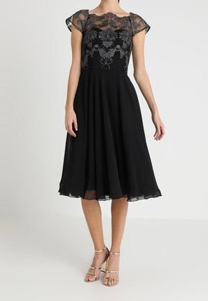 Woman wearing a black midi dress with sheer floral embroidered bodice and short sleeves, paired with gold strappy high-heeled sandals.
