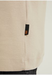 Helle beige Baumwoll-T-Shirt mit geradem Saum, ausgestattet mit einem seitlichen Etikett in Schwarz mit einem braunen quadratischen Akzent. Glatte Textur, einfaches Design.