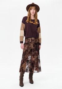 Bruine cropped trui met beige strepen, gelaagde bloemen kanten rok in bruine tinten, en bruine suède enkellaarsjes met hak, aangevuld met een bruine hoed.