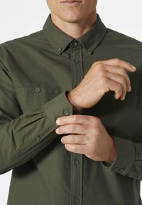 Camisa verde oliva de botones con cuello, dos bolsillos en el pecho y puños abotonados, con una textura suave y un diseño ajustado.