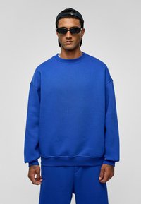 Blauwe fleece sweatshirt met ribbelstructuur, ronde halslijn en een relaxed fit. Gecombineerd met bijpassende shorts, gedragen met een zwarte pet en zonnebril.
