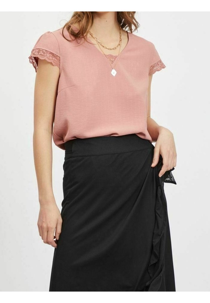 Femme portant un chemisier à manches courtes rose orné de dentelle, rentré dans une jupe noire taille haute avec un détail volanté sur le côté.