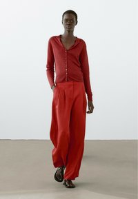 Rode gebreide cardigan met knopen aan de voorkant en capuchon, gecombineerd met wijde rode broek. Model draagt zwarte sandalen en staat op een neutrale vloer.