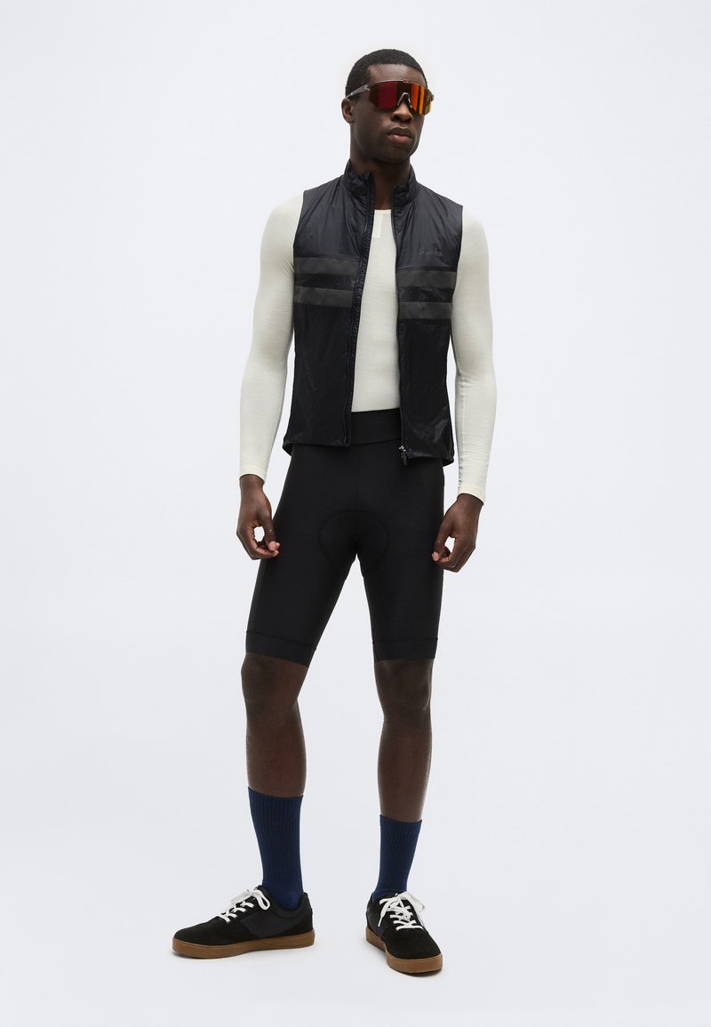 Man wearing black cycling shorts, white long-sleeve top, black sleeveless vest, blue socks, black sneakers, and red reflective sunglasses.