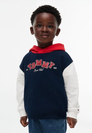 Jeune garçon portant un sweat à capuche Tommy Sport Club bleu marine, rouge et blanc et un jean bleu, souriant légèrement sur un fond blanc uni.