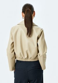 Veste courte beige en faux cuir lisse avec large col et taille élastique, assortie à un pantalon bleu marine à rayures.
