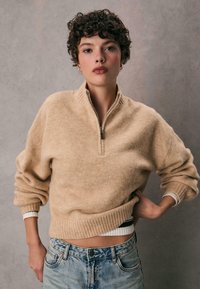 Beżowy sweter zapinany na zamek w miękkim dzianinowym materiale z prążkowanym wykończeniem u dołu i przy mankietach, zestawiony z jasnoniebieskimi dżinsami o klasycznym kroju.
