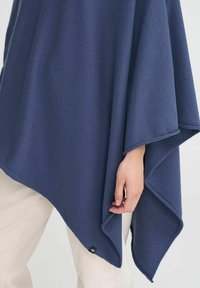 Mörkblå poncho i mjukt tyg med en asymmetrisk klippning och subtila sömmar. En liten etikett syns vid fållen.