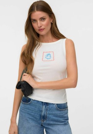 Vrouw met lang bruin haar die een witte mouwloze top met blauw schelppatroon en hooggetailleerde blauwe spijkerbroek draagt, en een kleine zwarte tas vasthoudt.