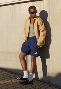 Yellow button-up shirt over a gray tank, paired with navy shorts featuring orange stripes. Black shoes, white socks, and a textured background.