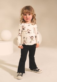 Haut à manches longues couleur crème avec imprimés floraux et personnages, associé à un pantalon évasé noir et des baskets noires à semelles blanches.