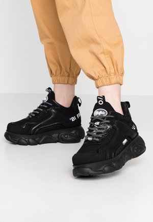 Henkilö, joka käyttää mustia paksupohjaisia lenkkareita, joissa on valkoiset merkit, ja taitettuja vaaleanruskeita housuja, seisoo yksinkertaisella vaalealla lattialla.